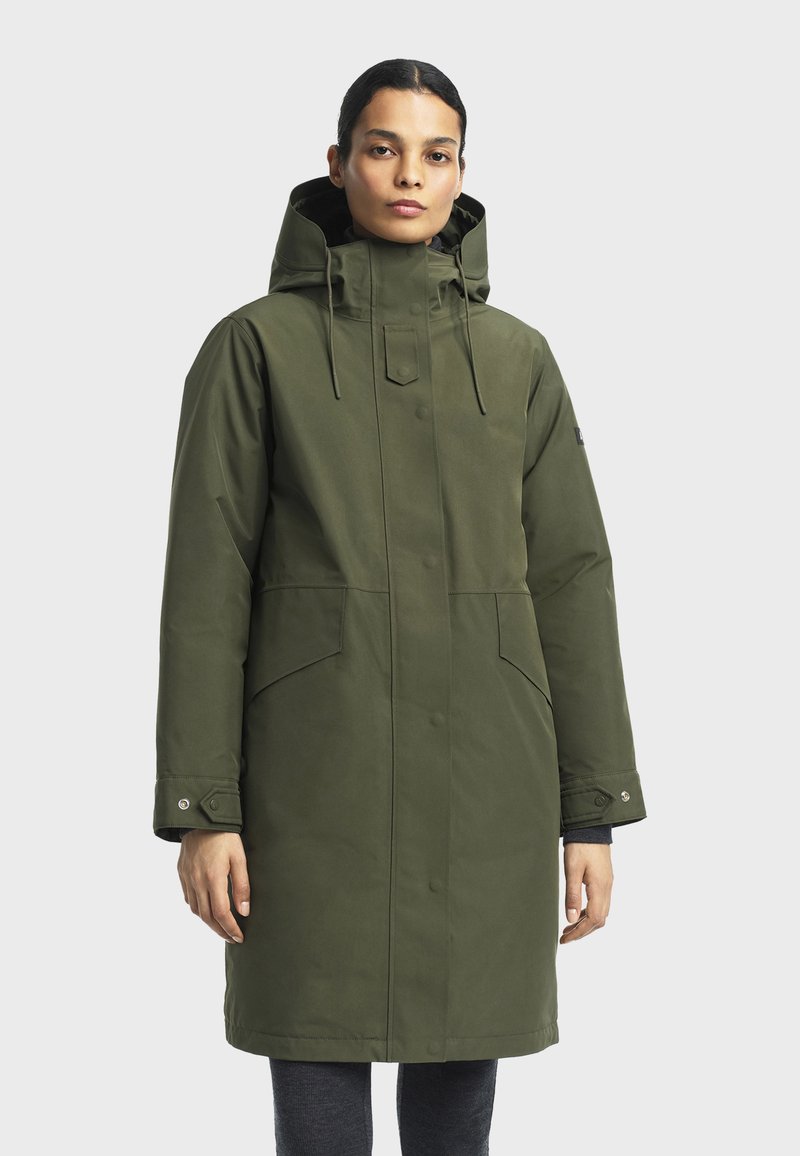 Parka imperméable vert olive avec capuche amovible, boutons-pression et deux poches latérales. Caractérisée par une coupe droite et une texture douce.
