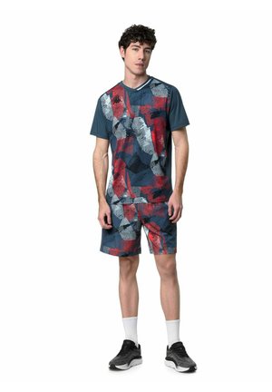 Giovane uomo con maglietta e pantaloncini sportivi coordinati con motivo astratto in blu navy, rosso e grigio, calzini bianchi e scarpe da ginnastica nere, in piedi.