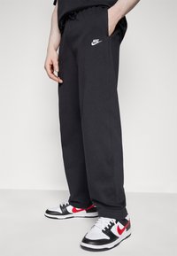 Uomo che indossa pantaloni di felpa neri Nike con sneaker bianche e rosse Nike, in piedi contro uno sfondo bianco uniforme.