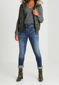 Gilet imbottito verde oliva con cappuccio rifinito in pelliccia sopra un maglione grigio, abbinato a jeans blu scuro con risvolto e stivaletti verde lucido.