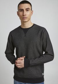 Sweatshirt noir avec un tissu texturé, col et poignets contrastés. Il présente un col rond, des manches longues et une petite étiquette de marque sur l'ourlet.