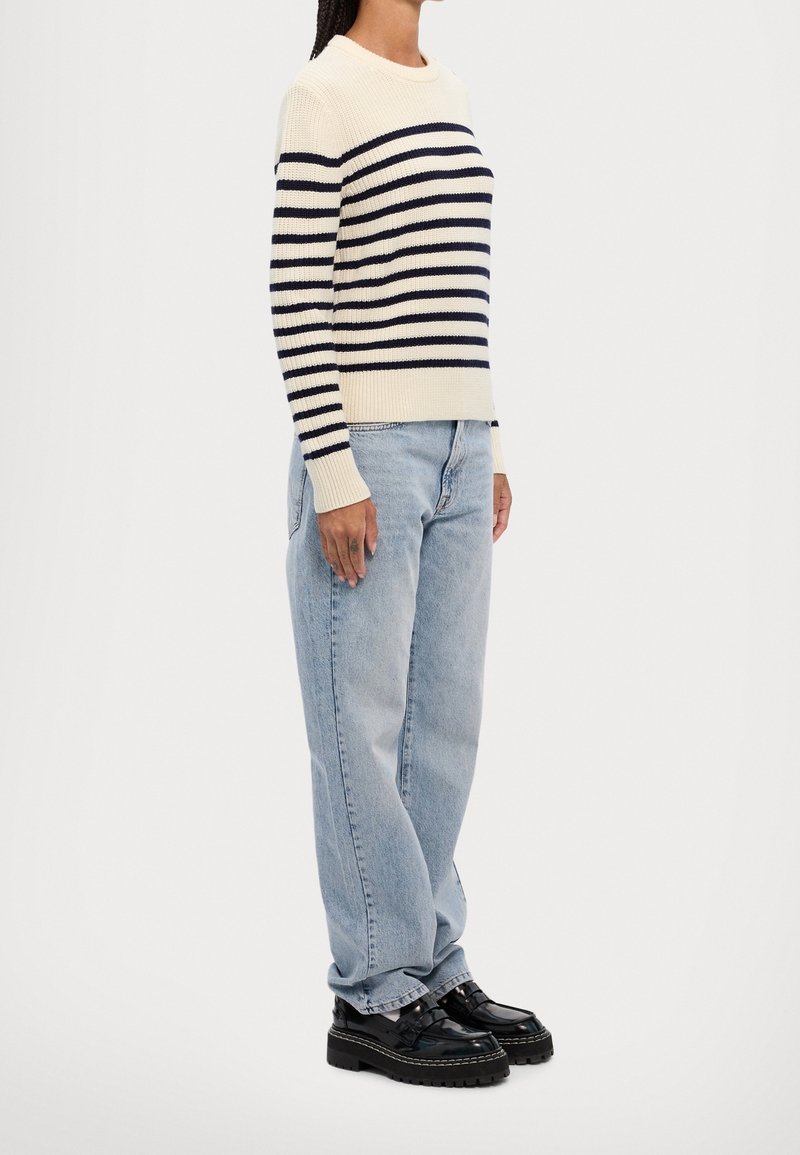Femme portant un pull crème à rayures horizontales noires, un jean taille haute bleu clair et des mocassins noirs épais, debout de côté.