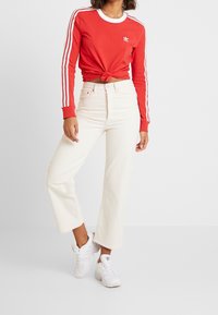 Röd långärmad skjorta med vit kant och tre vita ränder på ärmarna, knuten i midjan, tillsammans med högmidjade vita vida jeans och vita sneakers.