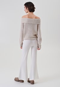 Donna con capelli corti e scuri, che indossa un maglione beige a spalle scoperte, pantaloni bianchi a palazzo e stivali marroni con tacco spesso, in piedi su uno sfondo neutro.