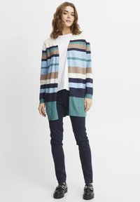 Svītrots džemperis gaiši zilā, tumši zilā un zemes toņu krāsās ar atvērtu priekšpusi, kabatām un zaļganu apdari, apvienots ar tumšiem biksēm un apaviem.