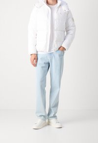 Osoba w białej kurtce puchowej, jasnoniebieskich dżinsach, białych sneakersach, z jedną ręką w kieszeni na tle jednolitego, jasnego tła.