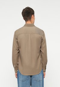 Cămașă maro cu nasturi, cu mâneci lungi și detaliu pe spate. Fabricată dintr-un material fin, cu tiv curbat și manșete cu nasturi.