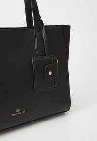 Geantă tote neagră din piele texturată, cu un buzunar cu fermoar atașat. Prezintă un logo auriu și un design minimalist.