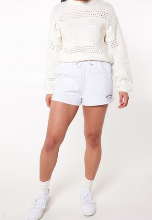 Maglione bianco a maglia con motivi a punto aperto, abbinato a pantaloni short in cotone grigi con logo. Sneakers bianche e calzini alla caviglia completano il look.