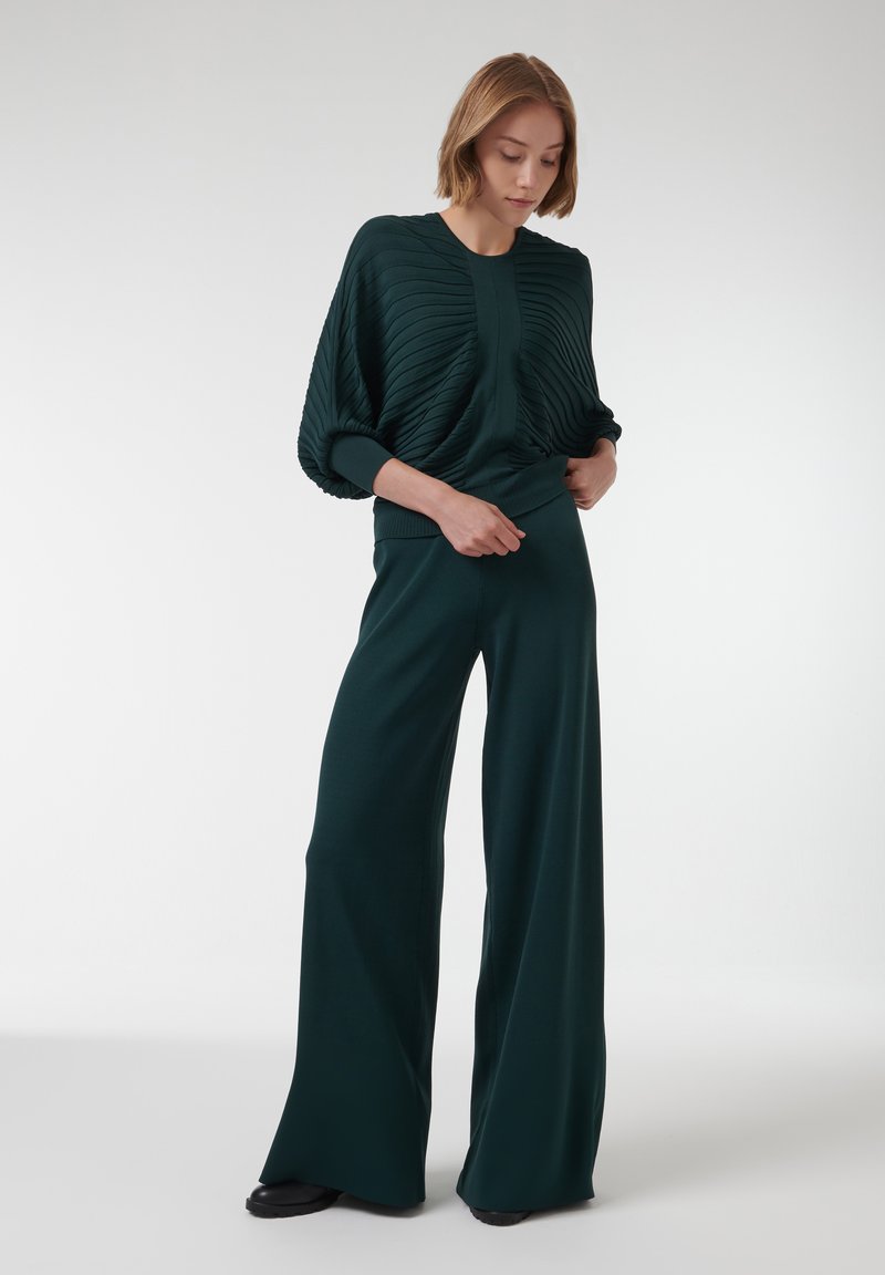 Diepgroene, geribbelde gebreide trui met een ontspannen pasvorm en wijde mouwen, gecombineerd met wijde broekspijpen van een vergelijkbaar getextureerd materiaal.