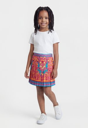 Jeune fille aux cheveux tressés portant un t-shirt blanc, une jupe à motifs colorés et des baskets blanches, debout et souriante sur un fond blanc.