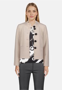 Beżowa, teksturowana kurtka z kołnierzem mandaryńskim, pięcioma czarnymi guzikami i dwiema przednimi kieszeniami, noszona na wzorzystej czarno-białej bluzce.