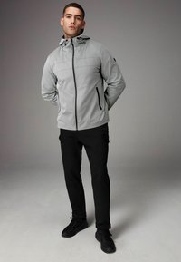 Jachetă zip-up de culoare gri deschis, cu glugă, având două buzunare laterale cu fermoar și accente negre contrastante. Purtați cu pantaloni negri și adidași.