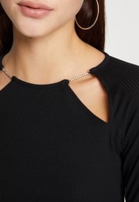 Svart ribbad topp med axelutskärningar, prydd med en silverkedja. Urrigningen är rundad och tyget har en texturerad yta.