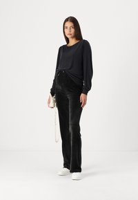 Schwarze Bluse mit langen Ärmeln und gerafften Ärmeln, kombiniert mit hoch taillierten, weichen schwarzen Cordhosen und weißen Sneakers. Weiße Schultertasche inklusive.