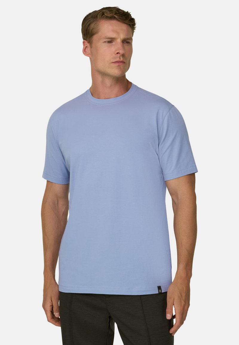 Homme portant un t-shirt bleu clair uni à manches courtes et un pantalon sombre, debout avec une expression neutre regardant légèrement sur le côté.