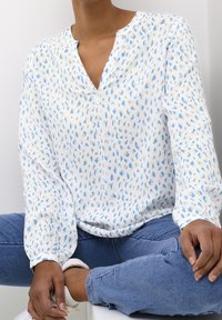 Blouse blanche avec imprimé abstrait bleu, tissu léger, encolure en V et manches longues. Associée à un jean bleu et des baskets blanches.