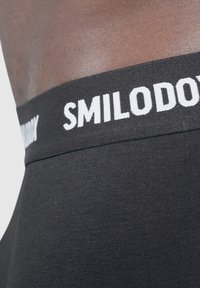 Černé spodní prádlo s hladkou texturou, které má široký elastický pas s nápisem "SMILODONY" v bílé barvě. Detailní záběr zvýrazňuje detaily šití.
