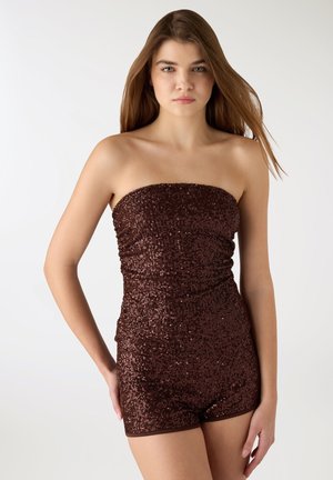 Giovane donna con capelli lisci castano chiaro che indossa una tuta senza spalline di paillettes marrone scuro, in piedi davanti a uno sfondo bianco semplice.