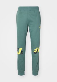 Green cotton sweatpants with a tapered fit, featuring large yellow and black graphic prints on the lower legs and an elastic waistband.