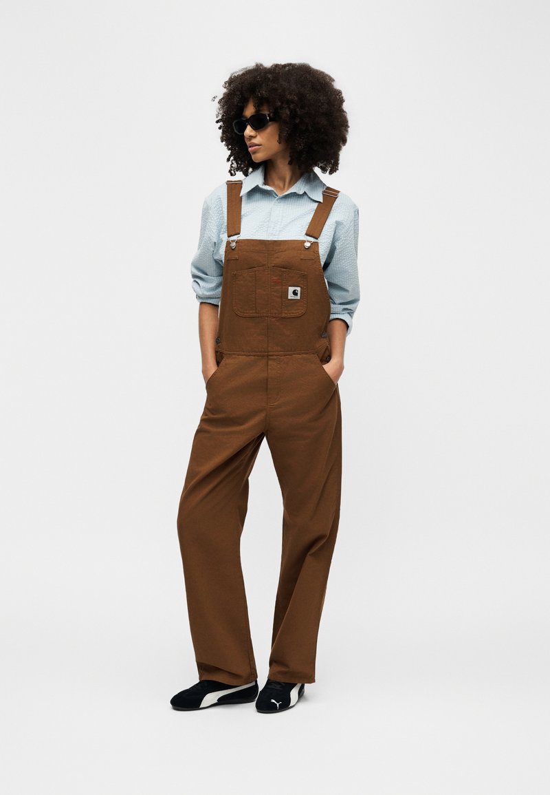 Woman with curly hair wearing black sunglasses, light blue shirt, brown overalls, and black Puma shoes, standing with hands in pockets.