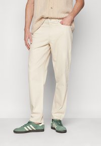 Pantalones de algodón beige con un corte holgado, que cuentan con bolsillos y una textura suave. Combinados con zapatillas de ante verde con rayas blancas.