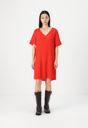 Robe rouge à manches courtes avec un décolleté en V profond, en tissu texturé et à coupe ample, associée à des bottes marron jusqu'aux genoux.
