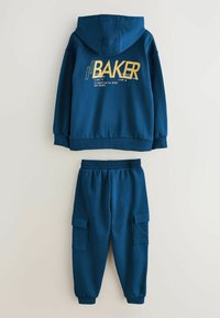 Marineblauwe hoodie met gele tekst en logo op de achterkant, gecombineerd met bijpassende joggerbroeken met cargozakken en elastische boorden.