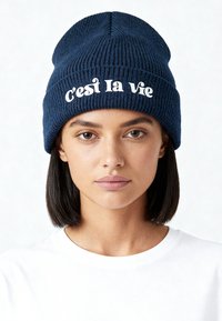 Jonge vrouw met recht donker haar, een marineblauwe gebreide muts met de tekst "c'est la vie" en een eenvoudige witte T-shirt, tegen een lichte achtergrond.