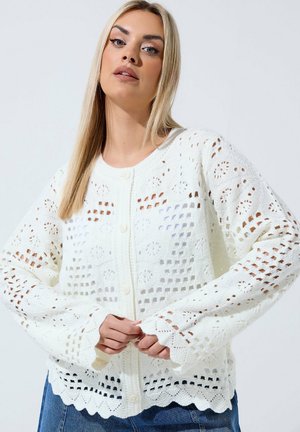 Kobieta z długimi blond włosami, ubrana w biały szydełkowany sweter na guziki i niebieskie dżinsy, poprawiająca rękawy na jednolitym tle.