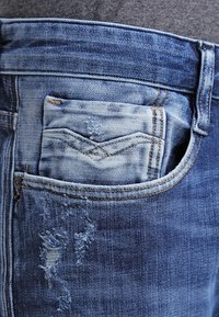 Blå denimjeans med en bakficka som har en urblekt plånbok. Tyget visar subtila slitningar och slitna fläckar, med kontrasterande sömmar.
