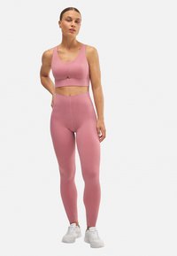 Sujetador deportivo de un suave color rosa claro con tirantes anchos, combinado con leggings de tiro alto y ajustados. Textura suave, costuras mínimas y sin patrones visibles.