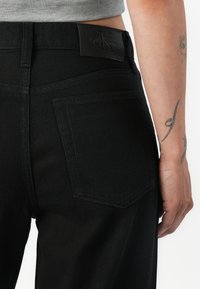 Svarta denimjeans med en bakficka, en läder CK-logotyppatch och en klassisk passform. Tyget har en slät textur med minimala sömdetaljer.
