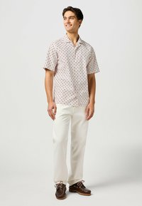 Camicetta a maniche corte di un rosa chiaro con motivi circolari, abbinata a pantaloni crema e scarpe nautiche marroni. Vestibilità slim, texture del tessuto liscia.