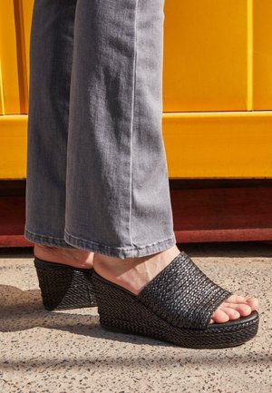 Sandalias de cuña tejidas en negro con un diseño texturizado. El zapato presenta una punta abierta y un tacón curvado, combinado con pantalones de pierna ancha en gris claro.