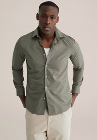 Lichtolijfgroen overhemd met knopen, lange mouwen, getailleerd ontwerp en witte knopen. Draagt het over een wit shirt, gecombineerd met beige broek.