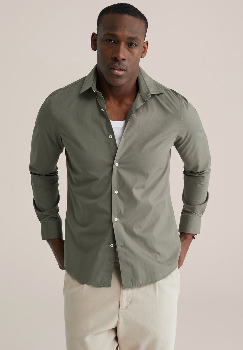 Lichtolijfgroen overhemd met knopen, lange mouwen, getailleerd ontwerp en witte knopen. Draagt het over een wit shirt, gecombineerd met beige broek.