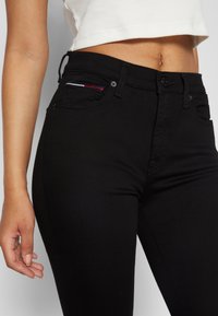 Jeans negros de talle alto con una textura suave. Presenta un bolsillo pequeño con costuras en rojo, blanco y azul. Diseño ajustado.