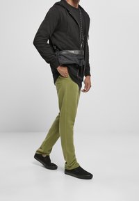 Sudadera negra con cremallera, pantalones de chándal verde oliva y zapatos negros sin cordones. El conjunto incluye un bolso negro tipo bandolera con una correa de cadena.