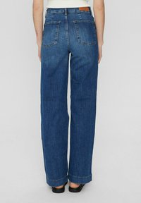 Breda jeans i mörkblå denim; har två bakfickor och en brun läderlapp vid midjan. Ren, rak skärning.