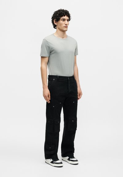 Νέος άνδρας με κατσαρά μαλλιά, φοράει ανοιχτόγκρι μπλουζάκι, μαύρο παντελόνι καμφορ με πολλές τσέπες και ασπρόμαυρα αθλητικά παπούτσια, στέκεται ακίνητος.