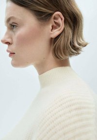 Maglione a coste color crema con colletto alto e dettagli texture, caratterizzato da una silhouette aderente e un tessuto morbido, posizionato su uno sfondo chiaro.