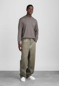 Pelēks garas piedurknes krekls ar mazu logotipu, apvienots ar brīvi pieguļošām olīvu zaļām biksēm un baltām kedām; gluda auduma un brīva piegriezuma.