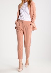 Svetle růžový oblek se zpevněným blejzrem a kalhotami. Pod ním bílá košile s knoflíky. Lesklé tělové lodičky doplňují outfit.
