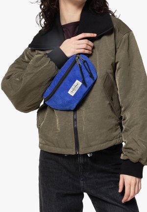Persoon draagt een olijfgroene bomberjas en zwarte broek, houdt een blauwe corduroy Eastpak heuptas over de borst.