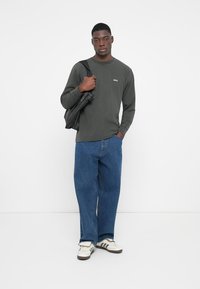 Langermet mørkegrønn skjorte med logo, kombinert med løse blå jeans og hvite joggesko med svarte detaljer. Modellen holder en svart veske.