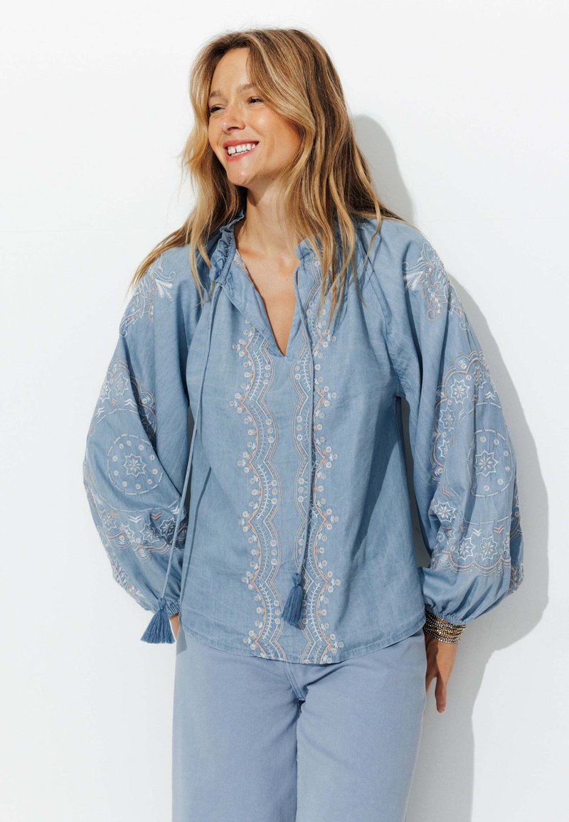 Blouse en coton bleu clair avec des manches ballon, des motifs floraux et géométriques brodés, un col à cordon et des accents de pompons.