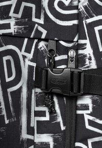 Svart tyg med djärv vit text och abstrakta mönster; detaljerad med en svart spänne, blixtlås och vävda remmönster.