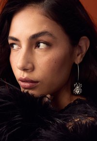 Silver spider-shaped hoop earring with intricate detailing, worn by a person against a dark background and soft black fur texture.