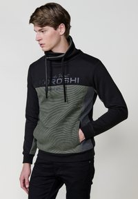 Sudadera negra y verde oliva con cuello alto, frente texturizado y detalles de logo. Presenta un ajuste relajado y mangas negras.